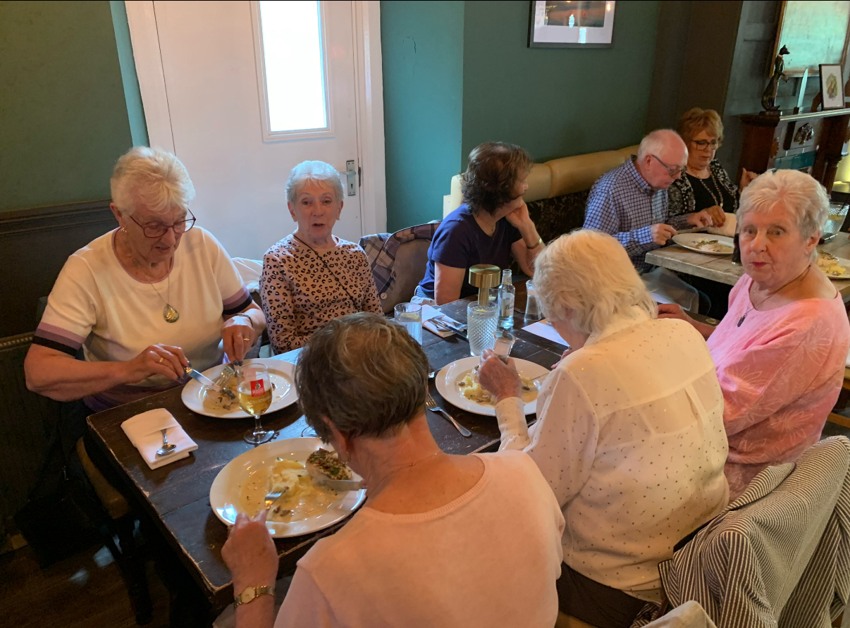 IMG_6424 6 ladies sitting at the dinner table eating their lunch off white plates, 2 ladies looking at the camera, a bit startled. A man and a lady in the background eating their lunch on another table